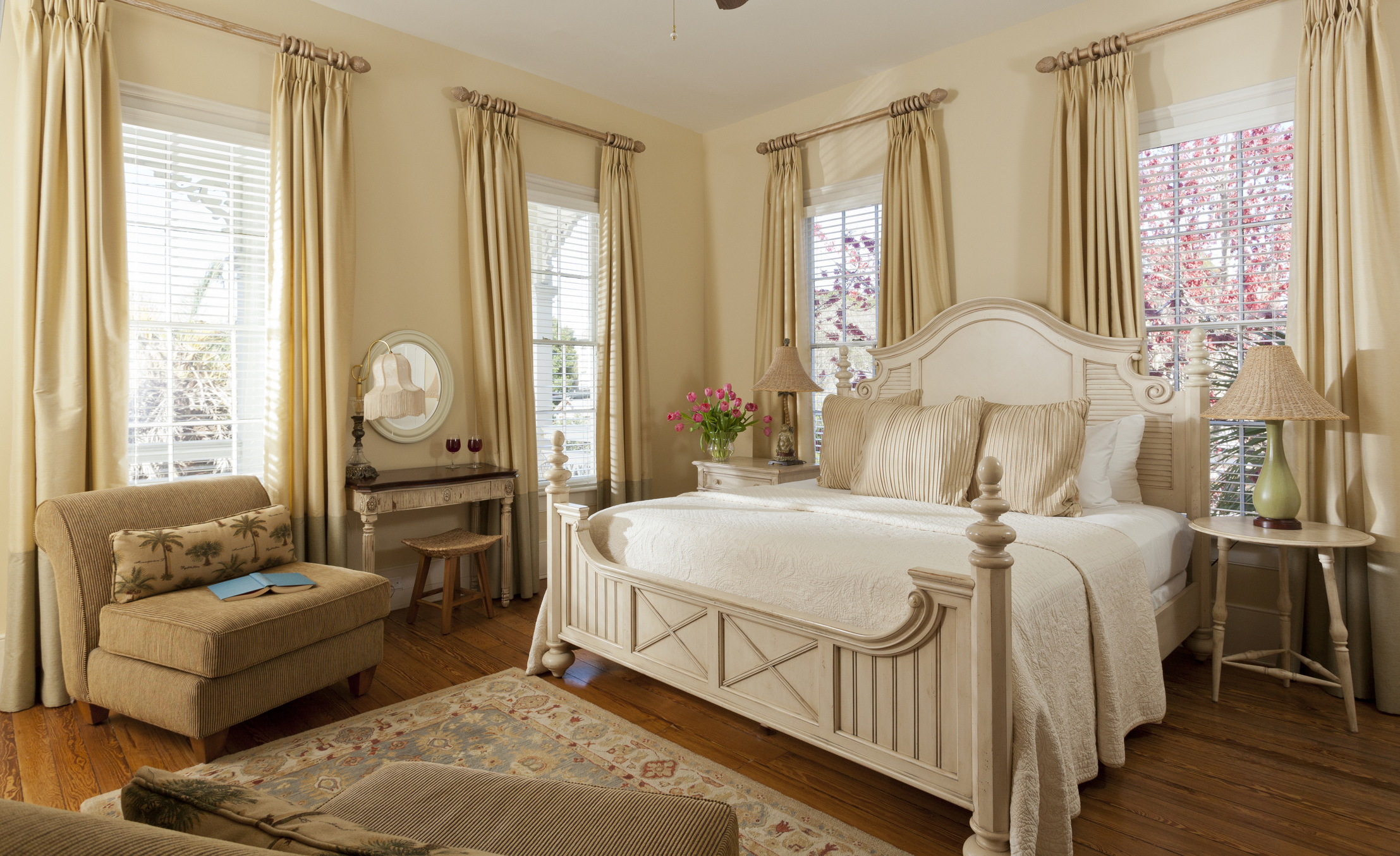 A beautiful guest bedroom replete with windows, two easy chairs, bedside tables, and a console table that doubles as a desk.