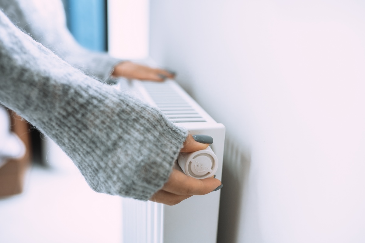 A woman with a gray sweater adjusts an older dial-styled thermostat.