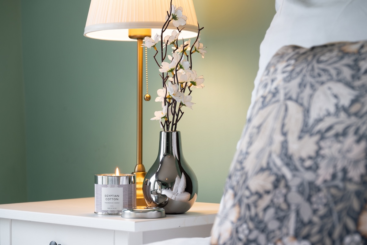 A bedside table with a lamp, silver vase, and a candle.