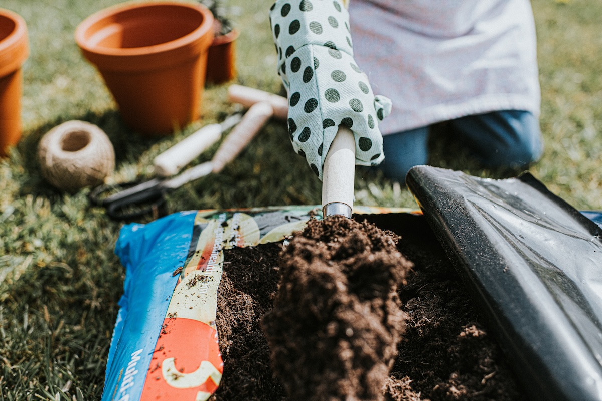 A gloved hand uses a garden shovel to open up a bag of potting soil.