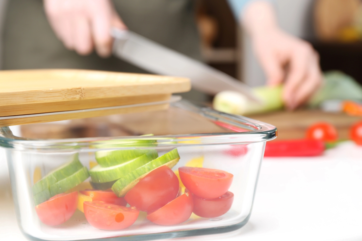 Glass container filled with cut vegetables with a woman cutting a leek in the background.