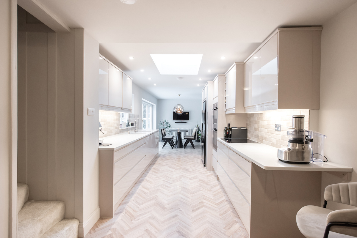 A clean white galley kitchen with white parquet flooring has a black dining table set at the end of it.