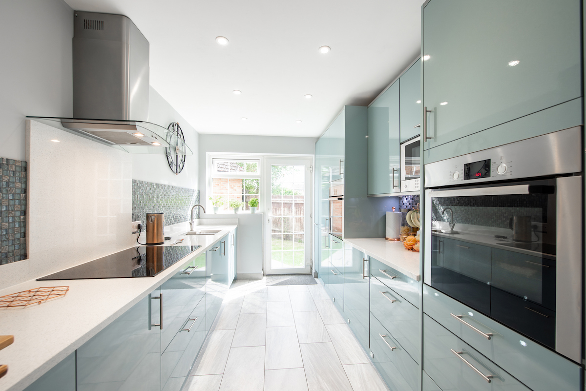 A galley kitchen with shiny mint green cabinets features a large window letting in natural light.