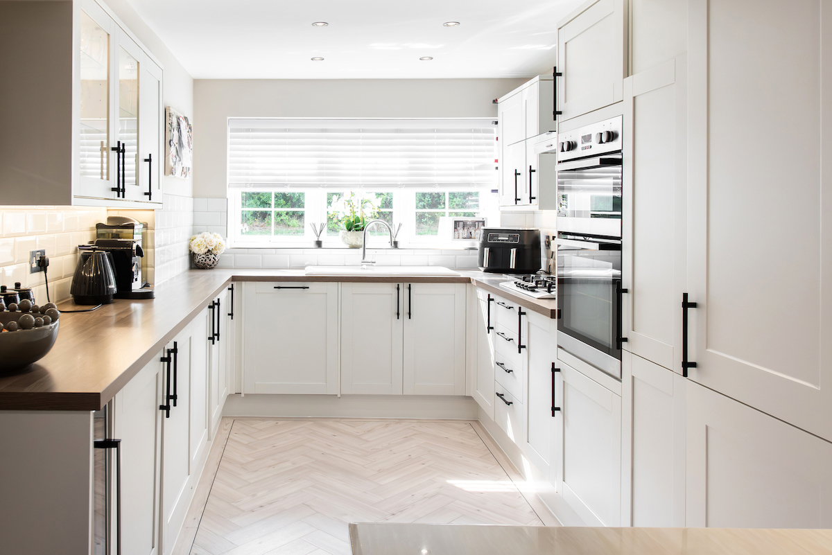 A galley kitchen with a large window is mostly white with black lower cabinet handles.