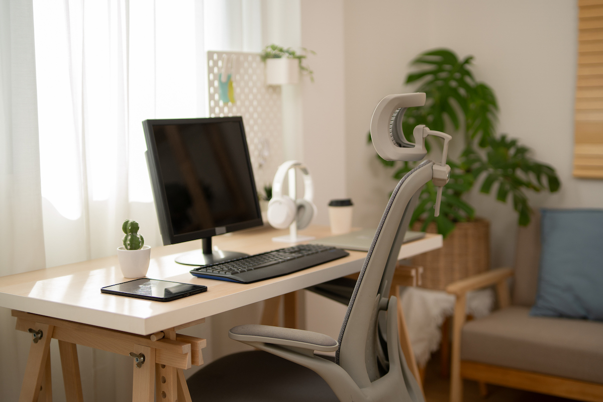 A home office desk, with a computer monitor and keyboard on top, and a desk chair are in front of a windows with sheer curtains.