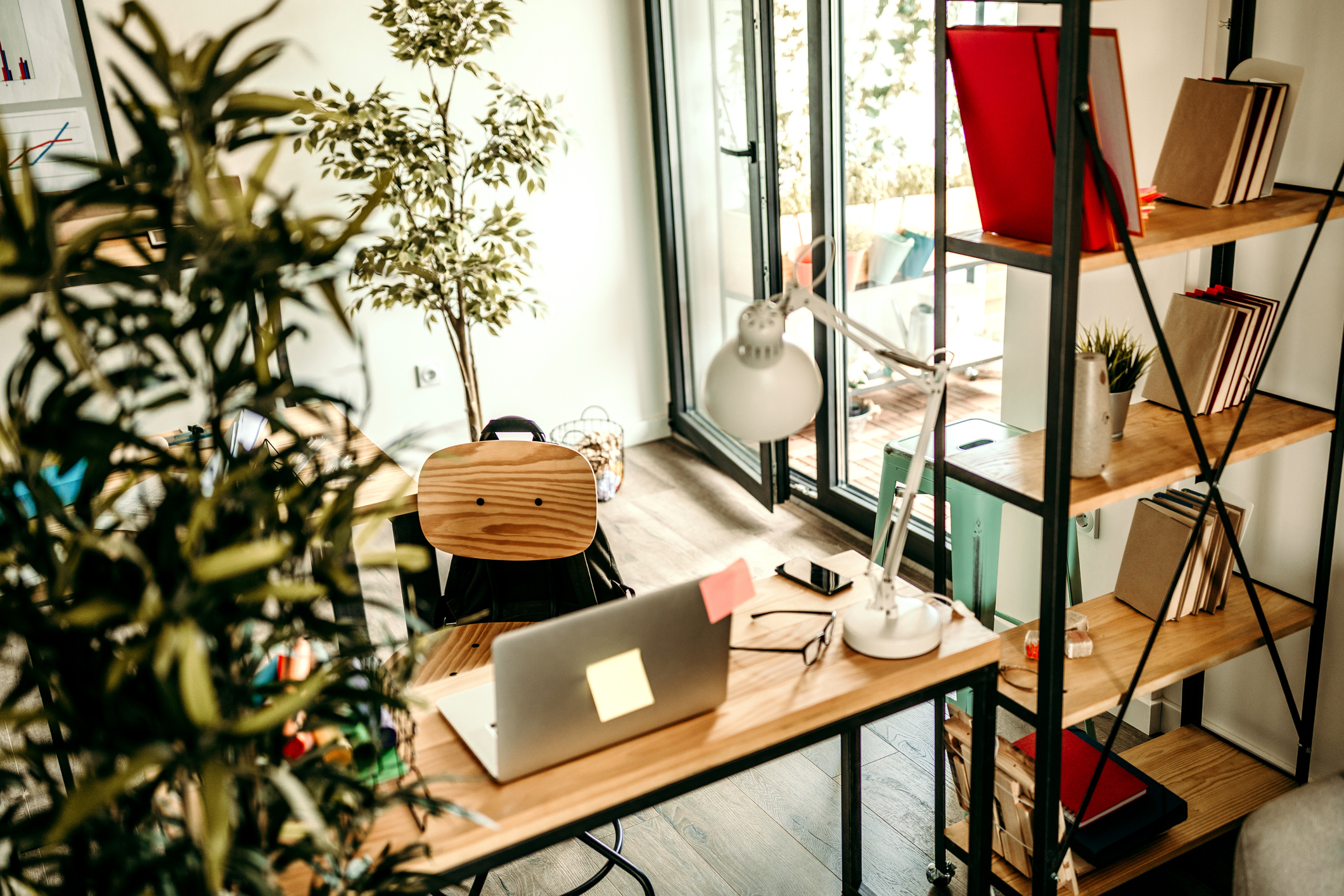 A small home office in a larger open-plan space, separated from the rest of the room with an open bookcase, simple desk, and large houseplant.