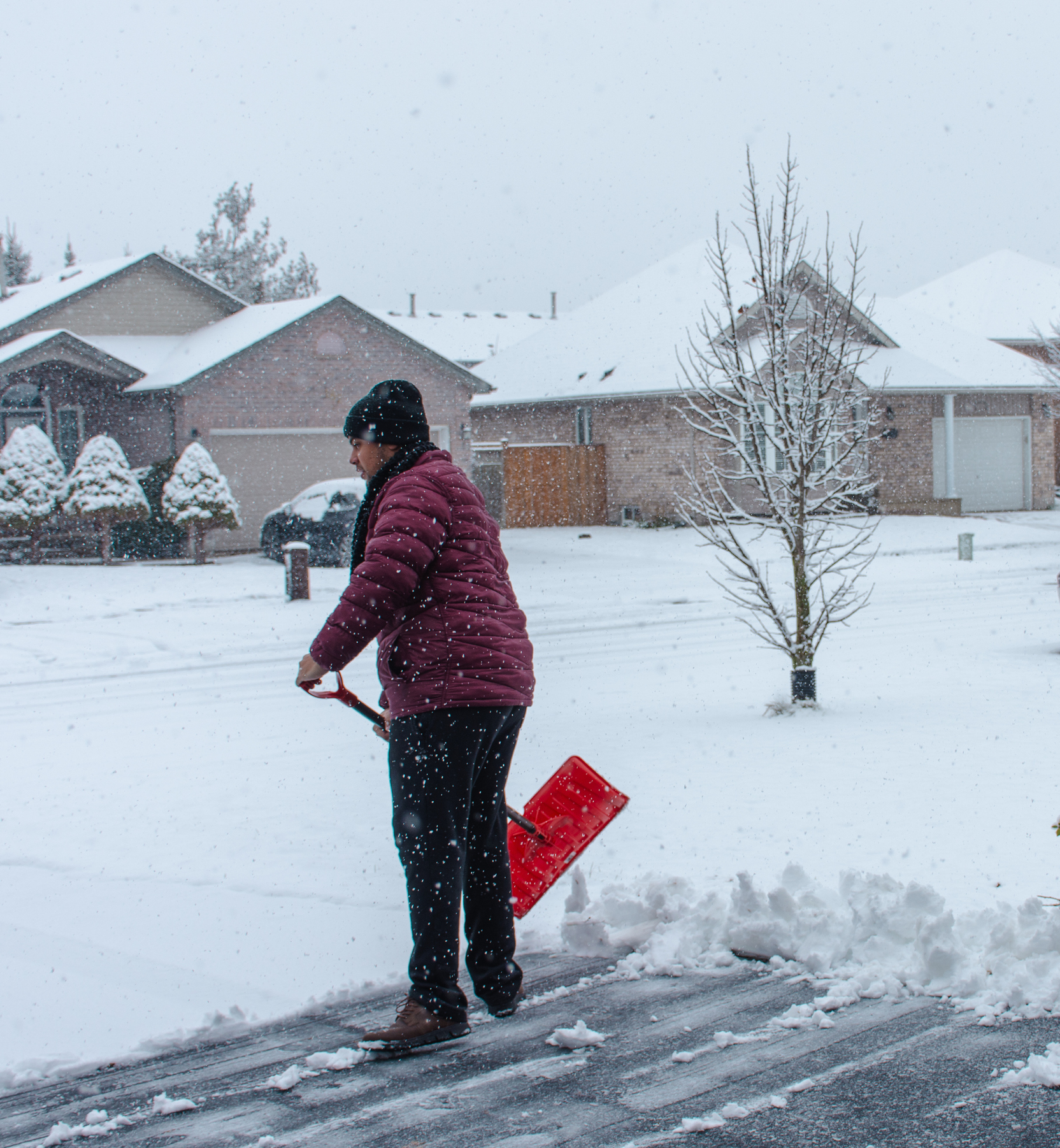 A man wearing layers of winter clothes shovels a snow-covered suburban driveway.