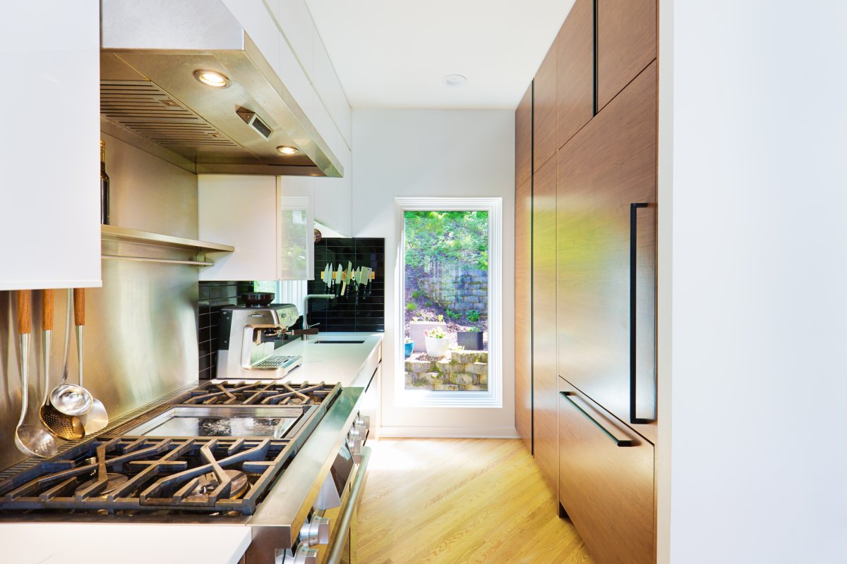 A sleek, modern galley kitchen with a walnut refrigerator built into matching ceiling-high cabinetry on the right wall and a professional cooking area on the left.