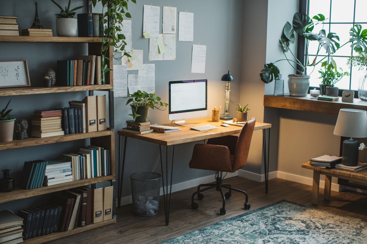 A modern home office with an open pine desk and bookshelf takes up a small corner of a larger room.