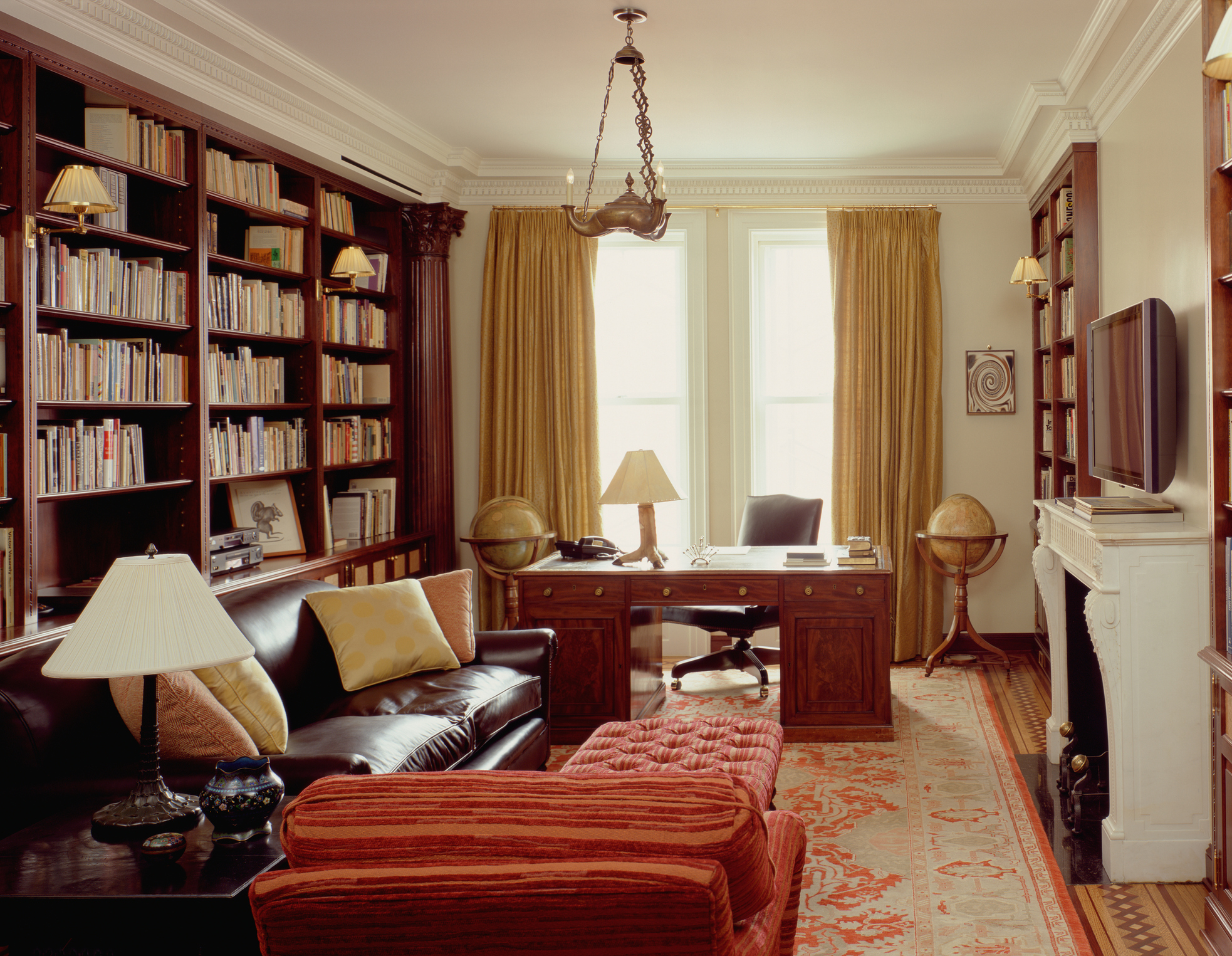 A warm home office with a large cherry wood desk in front of a window facing the rest of the furnished room.