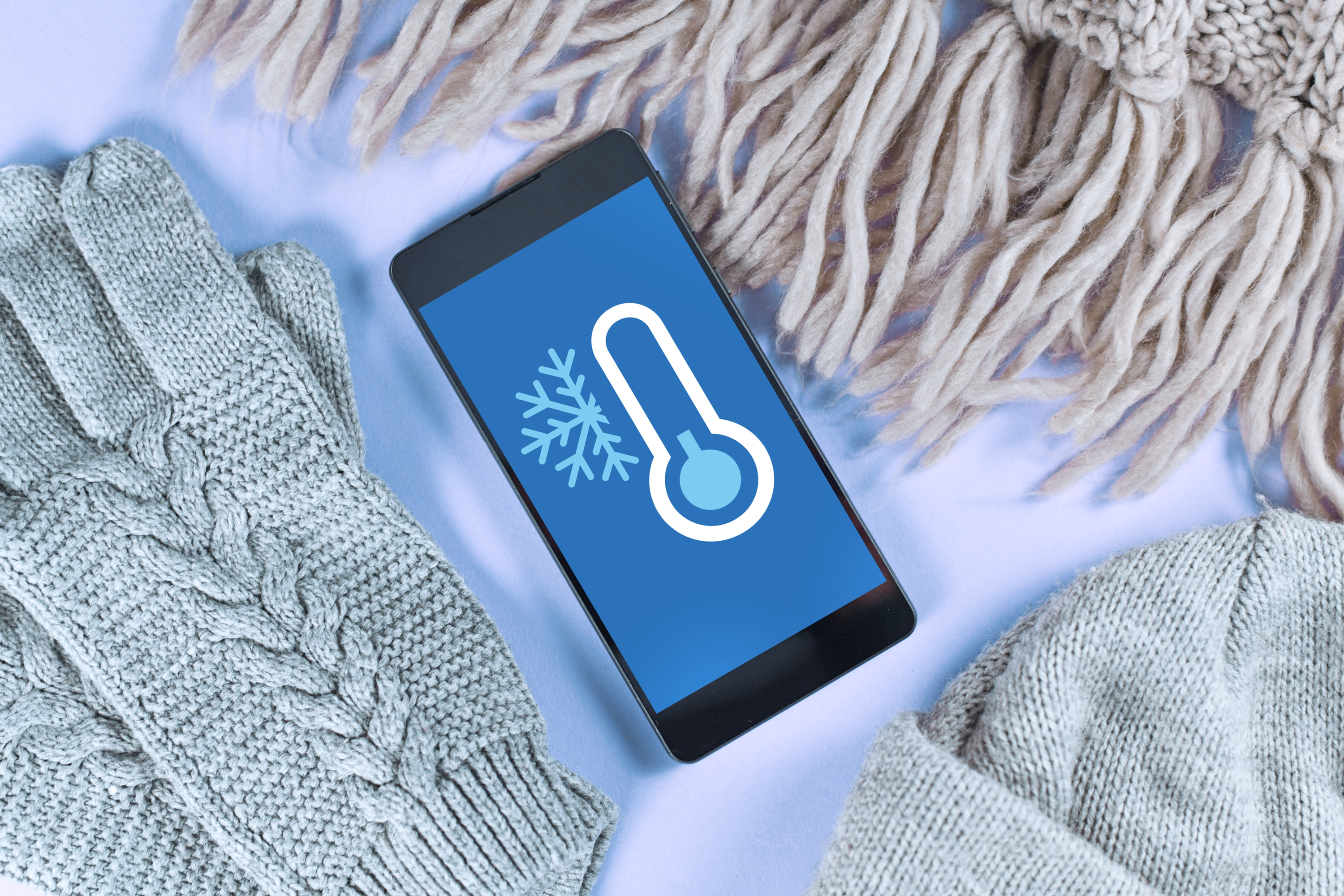 An iPhone with a screen-simulated winter weather related-image on its screen sits atop snow surrounded by winter gloves, a beanie, and scarf.