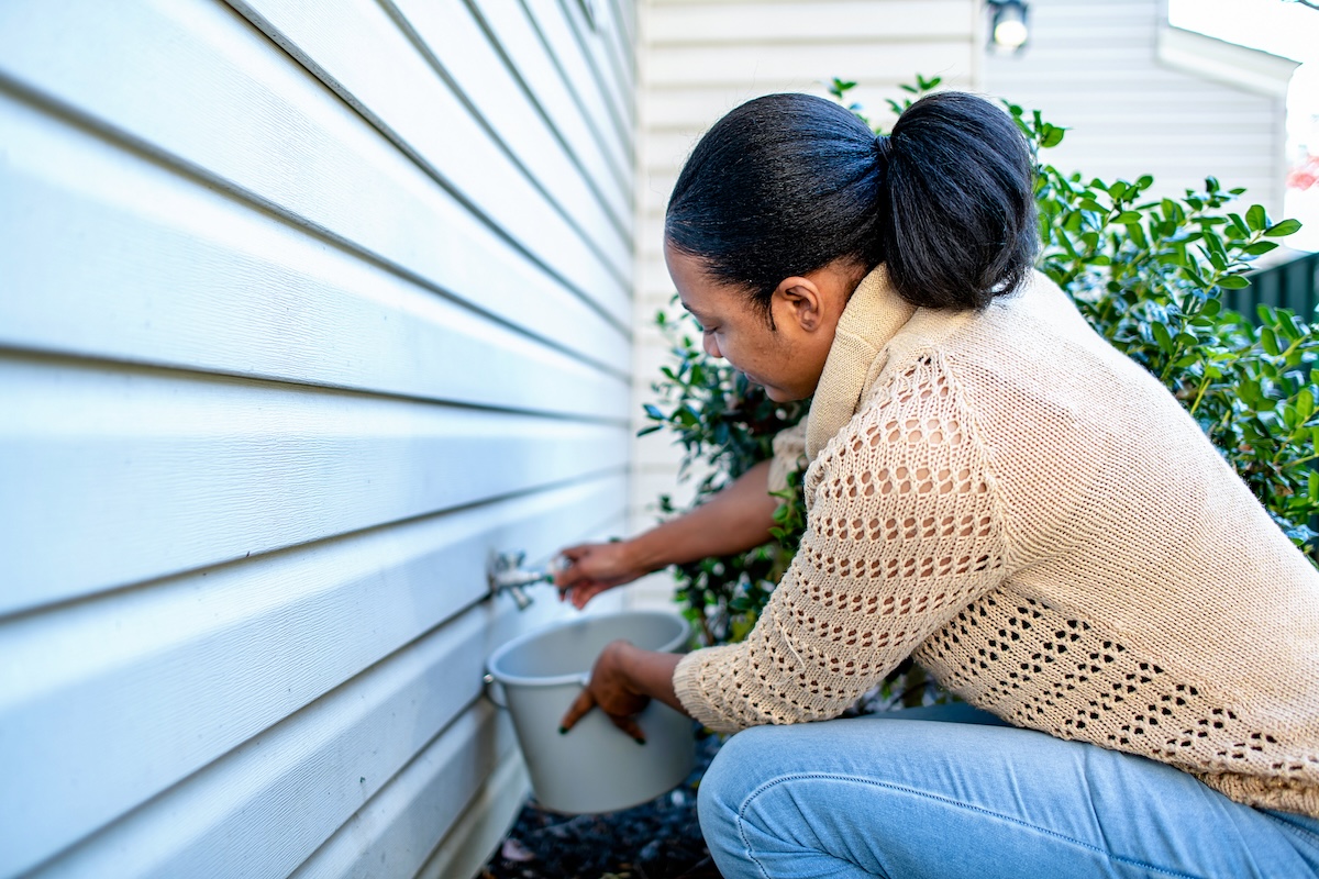 A woman in a sweater drains water from her home's exterior spigot.