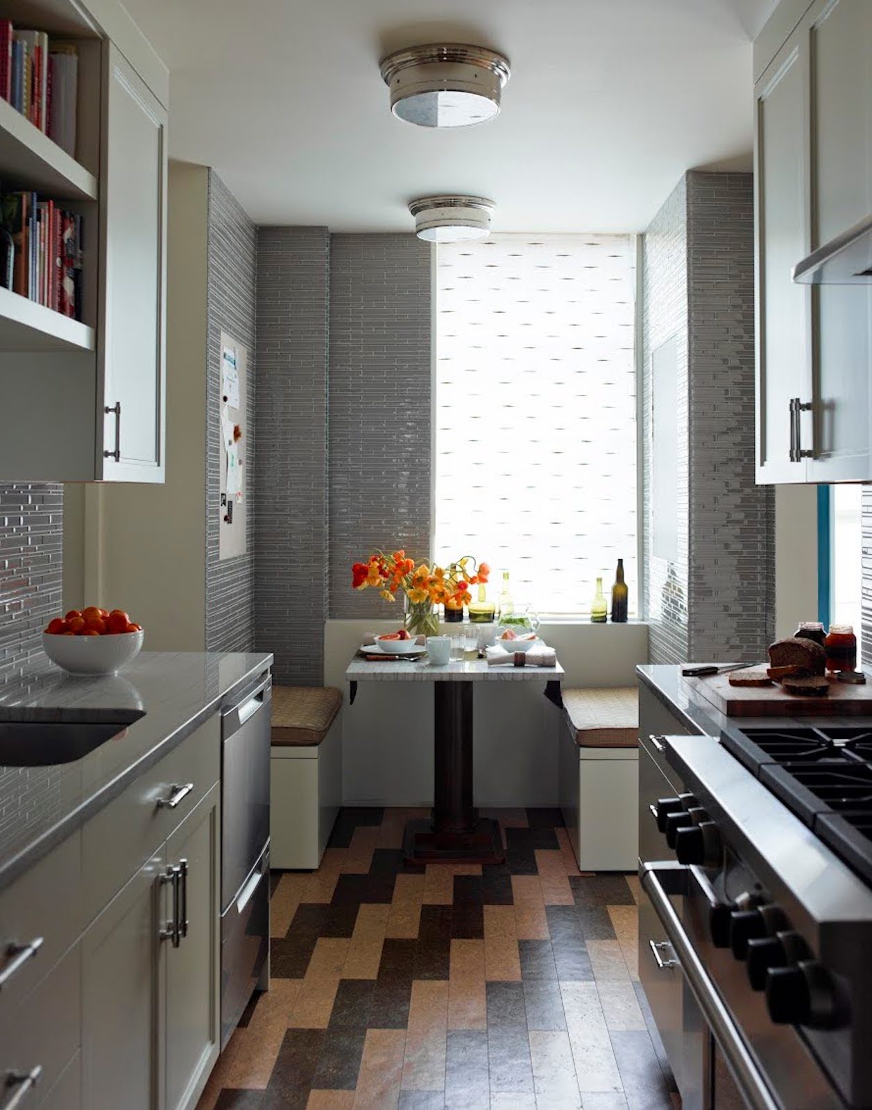 A galley kitchen with a large window features a wooden floor with alternating wooden and red colors.