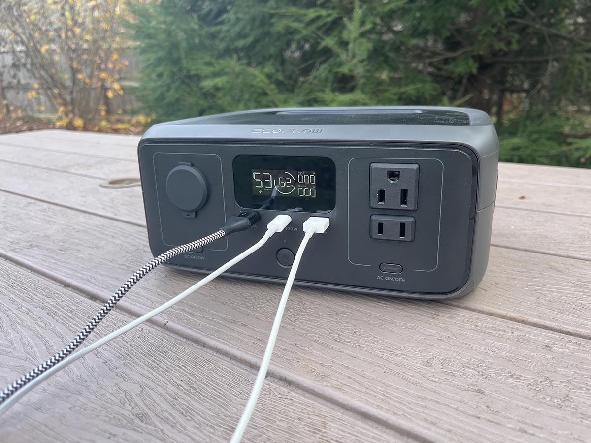 Three device charging cords plugged into the ports of the EcoFlow River 3 on a picnic table outside