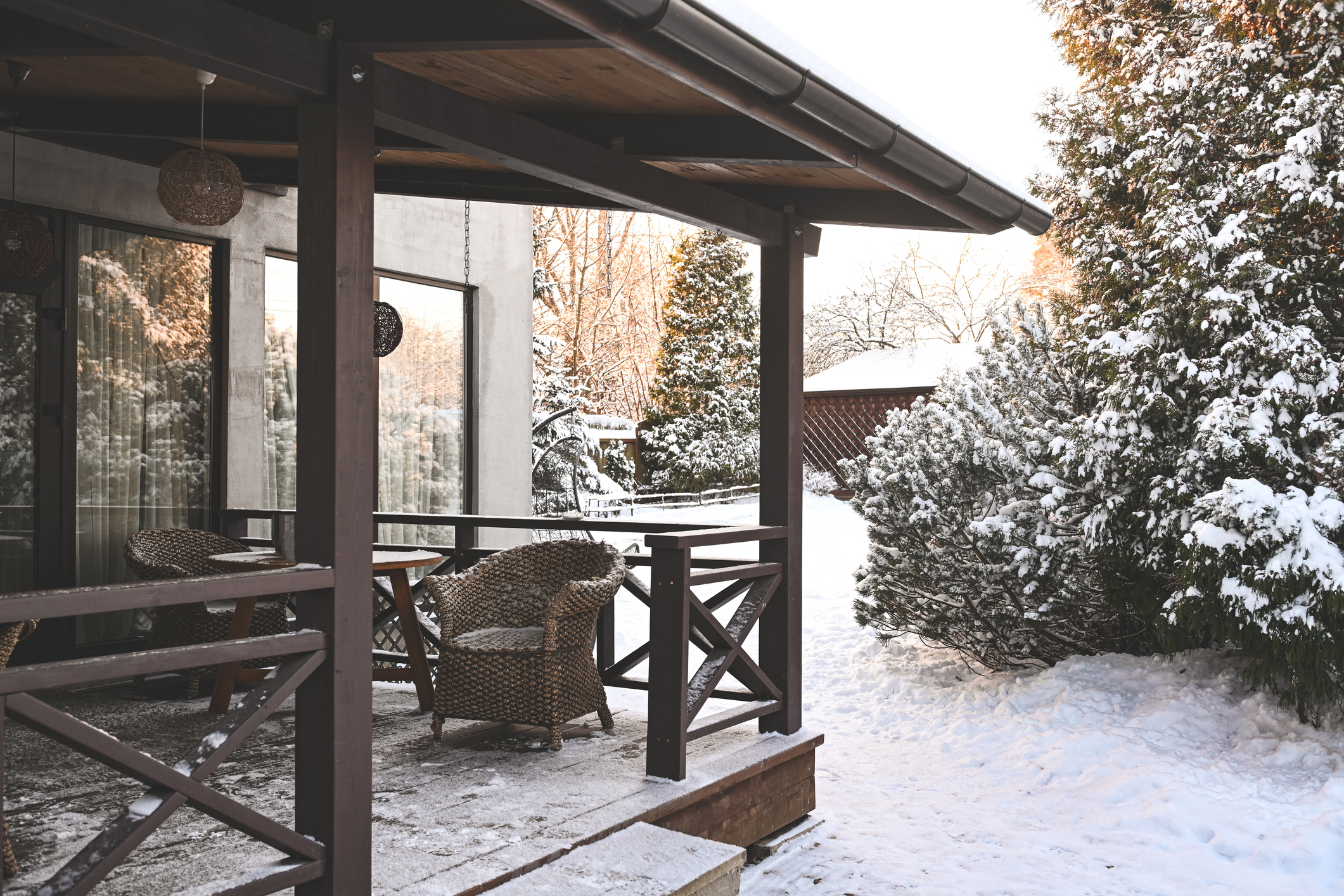 Snow-covered wooden house deck with trees in the background.