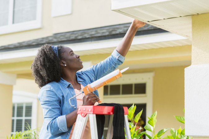 A homeowner on a ladder caulking gutters on a house.