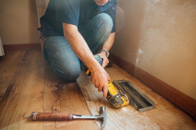 A man in jeans using an oscillating tool on an air vent embedded in wooden floors.