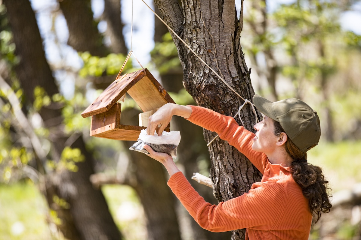 A woman in an orange jacket and green hat fills up a bird feeder on a sunny day.