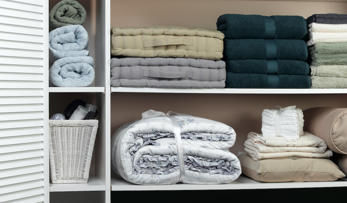 An open linen closet showing organized shelves of towels, cushions, sheets, and other linens of various colors.