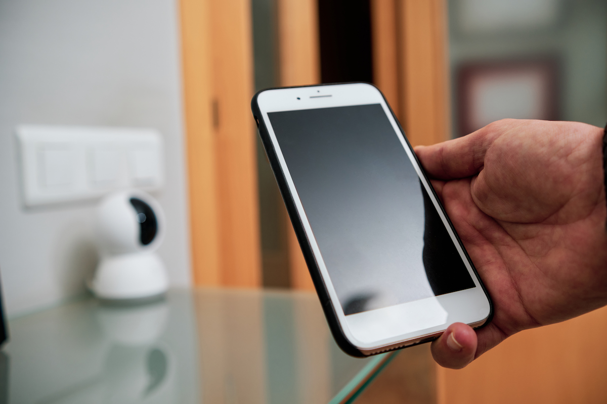 In foreground, man holds a smartphone in his hand; a home security camera sits on a table in the background.