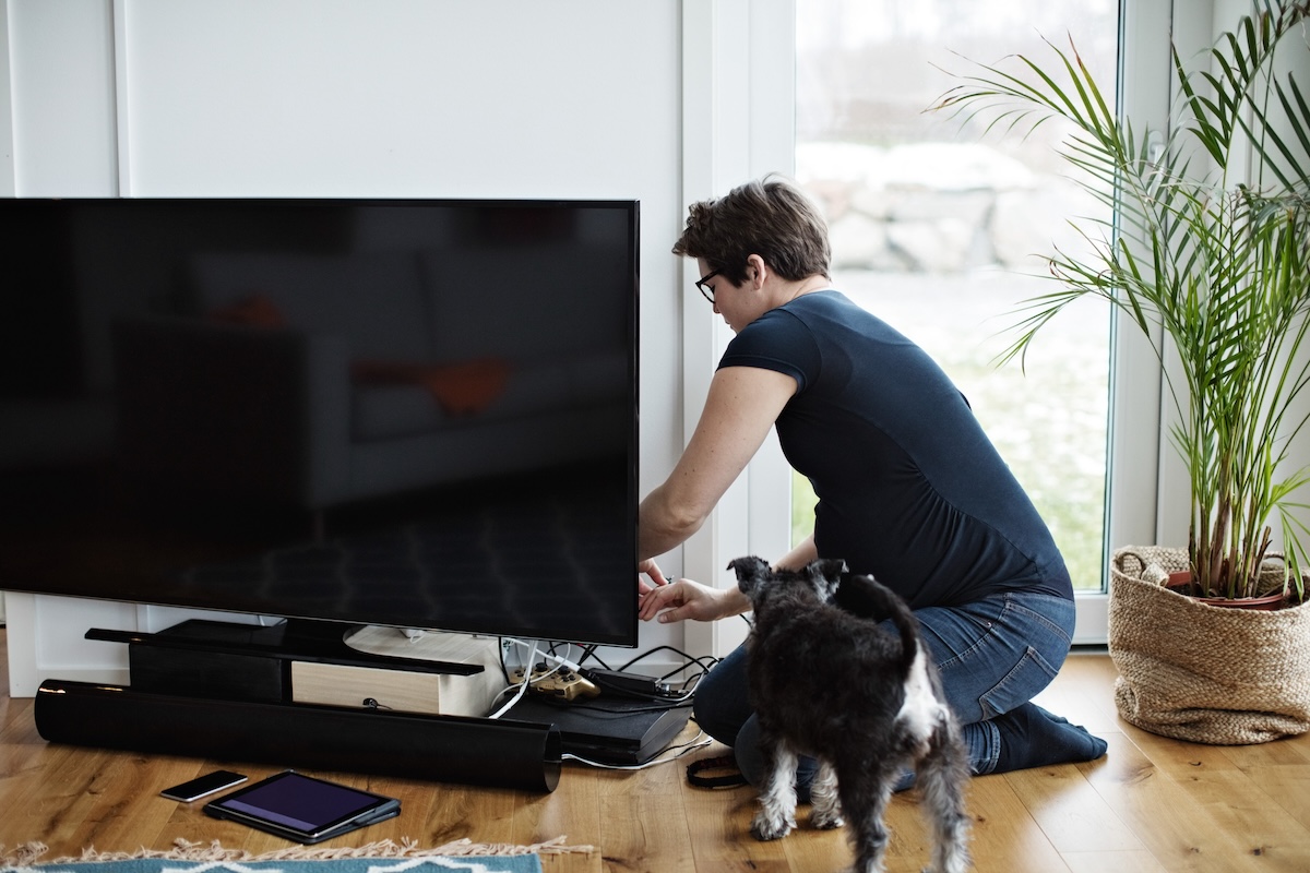 A woman with short hair hides TV wires behind a console as a dog looks on.