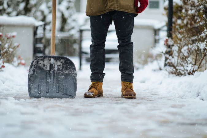 A homeowner dressed in winter clothes holding a shovel to clear a driveway of ice and snow.