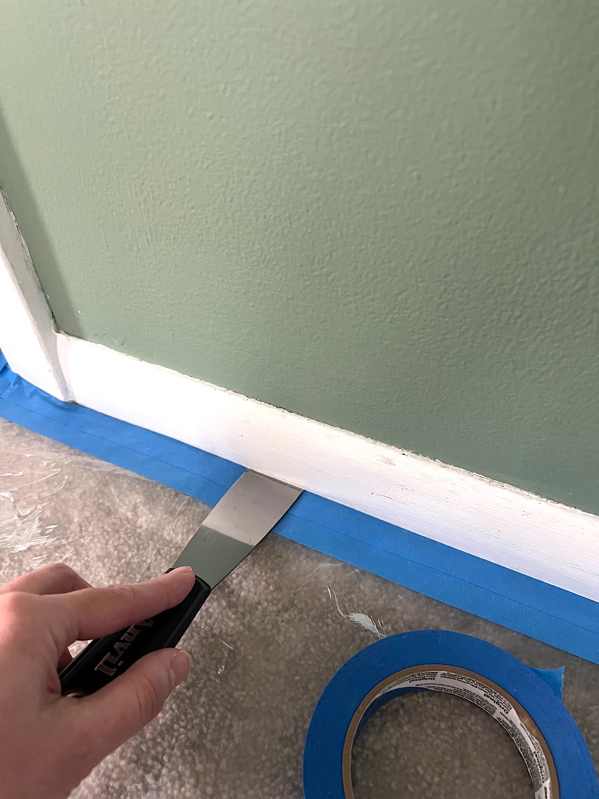 A DIYer using a putty knife to mask off carpet for painting baseboard trim.