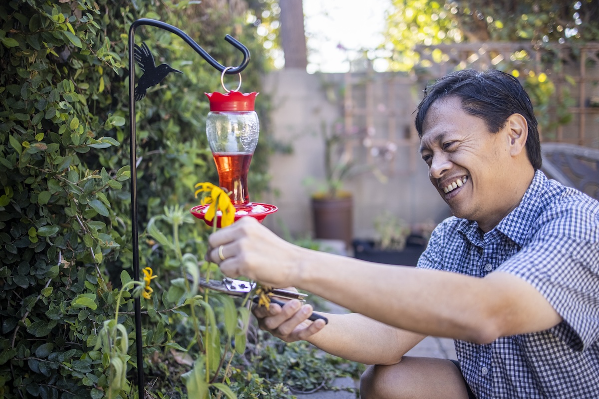 A man in his garden fixes a bright red hummingbird feeder.