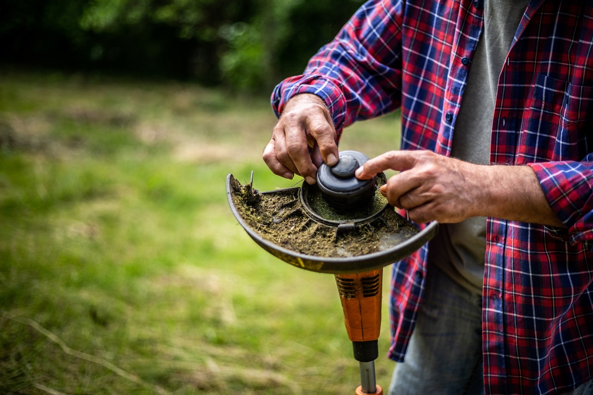 A man hold a weed eater machine upside down to remove the top.