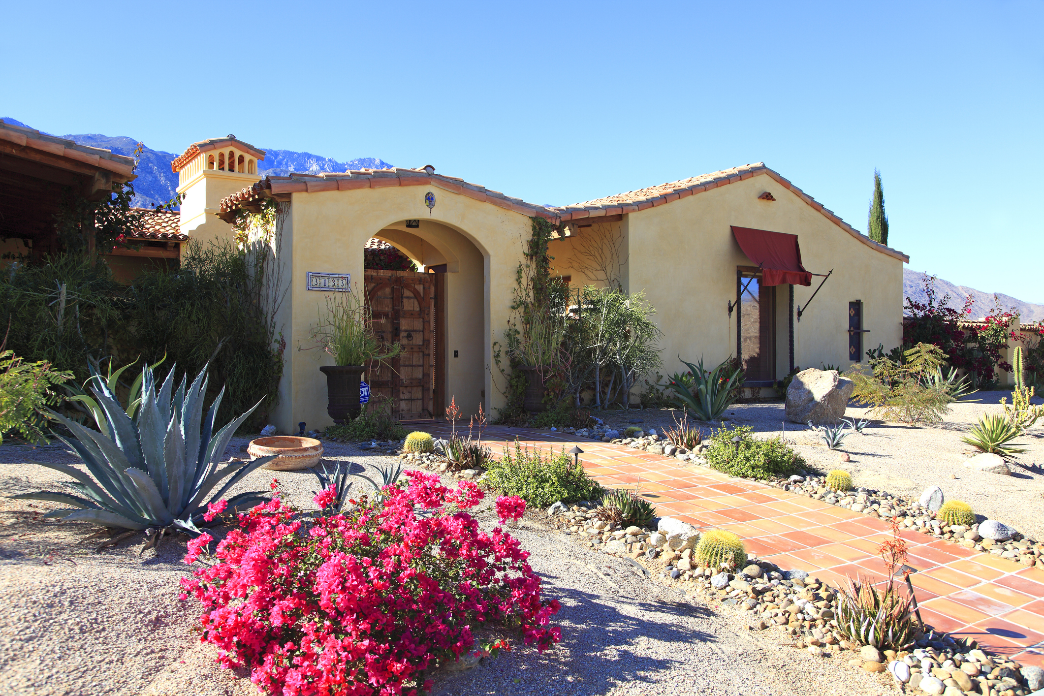 Tan adobe style home with simple but beautiful landscaping. Bougainvillea and cactus. Desert with Mountain range in background.